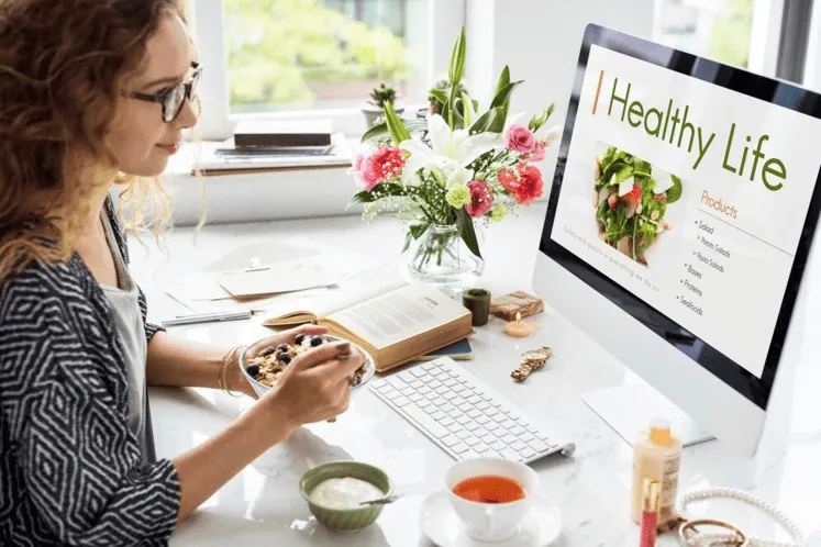 Person at a desk with a computer displaying 'Healthy Life,' a book, snack bowl, tea, and cosmetics.