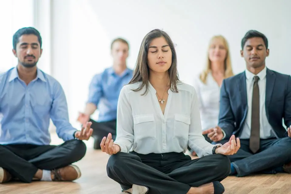Group of people sitting cross-legged and meditating in a bright office setting.