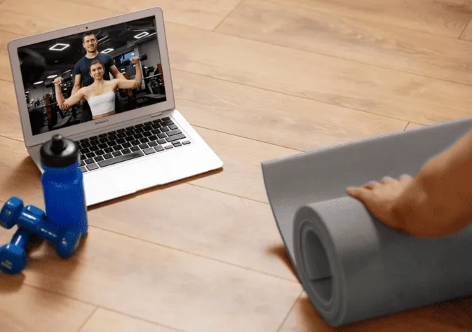 A laptop showing a workout video, a rolled-up exercise mat, and dumbbells on a wooden floor.s