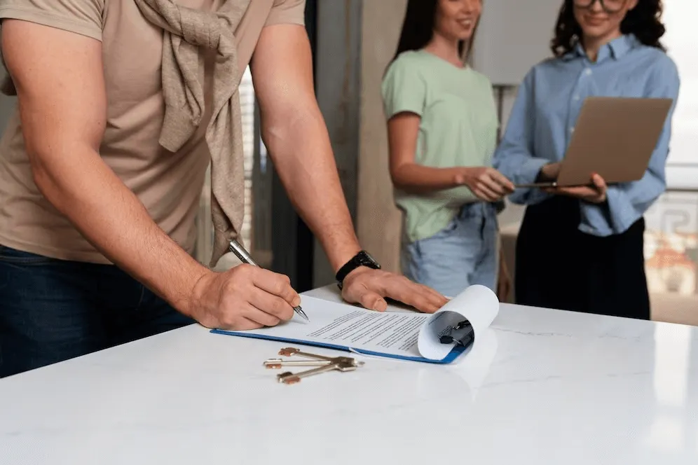 setting up a company in dubai - Person signing document on a table with keys beside, two observers standing with a laptop.