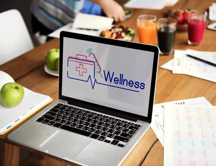 Mainland- Laptop on a wooden table displaying a 'Wellness' logo, surrounded by healthy food and drinks. 