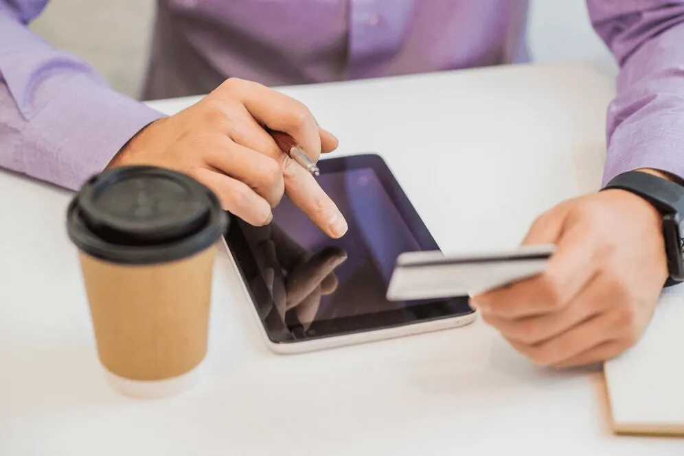 creating a company in dubai- Person using a stylus on a tablet next to a coffee cup and holding a card.